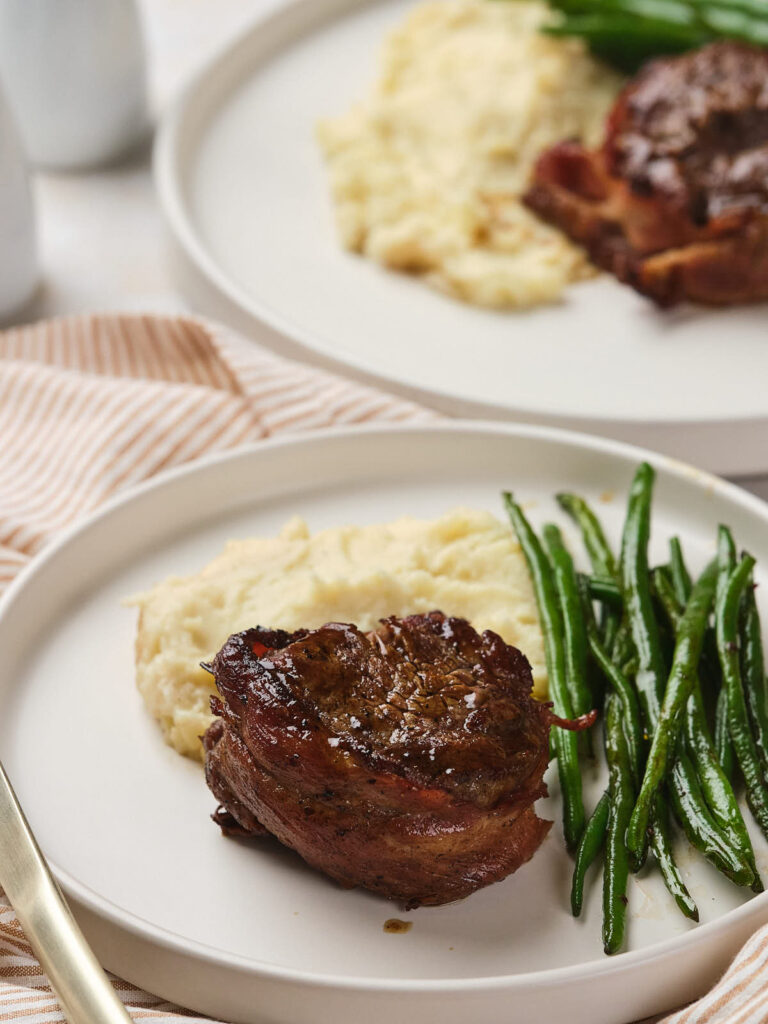 Plate with a bacon-wrapped filet mignon, mashed potatoes, and green beans, set on a striped napkin.