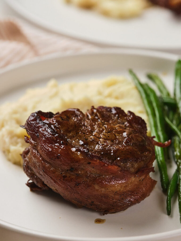 Steak wrapped in bacon served with mashed potatoes and green beans on a white plate.