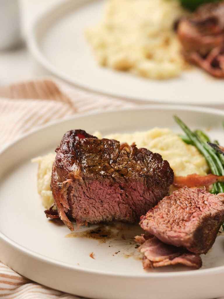 Steak with a slice cut out, served with mashed potatoes, greens, and bacon on a white plate.