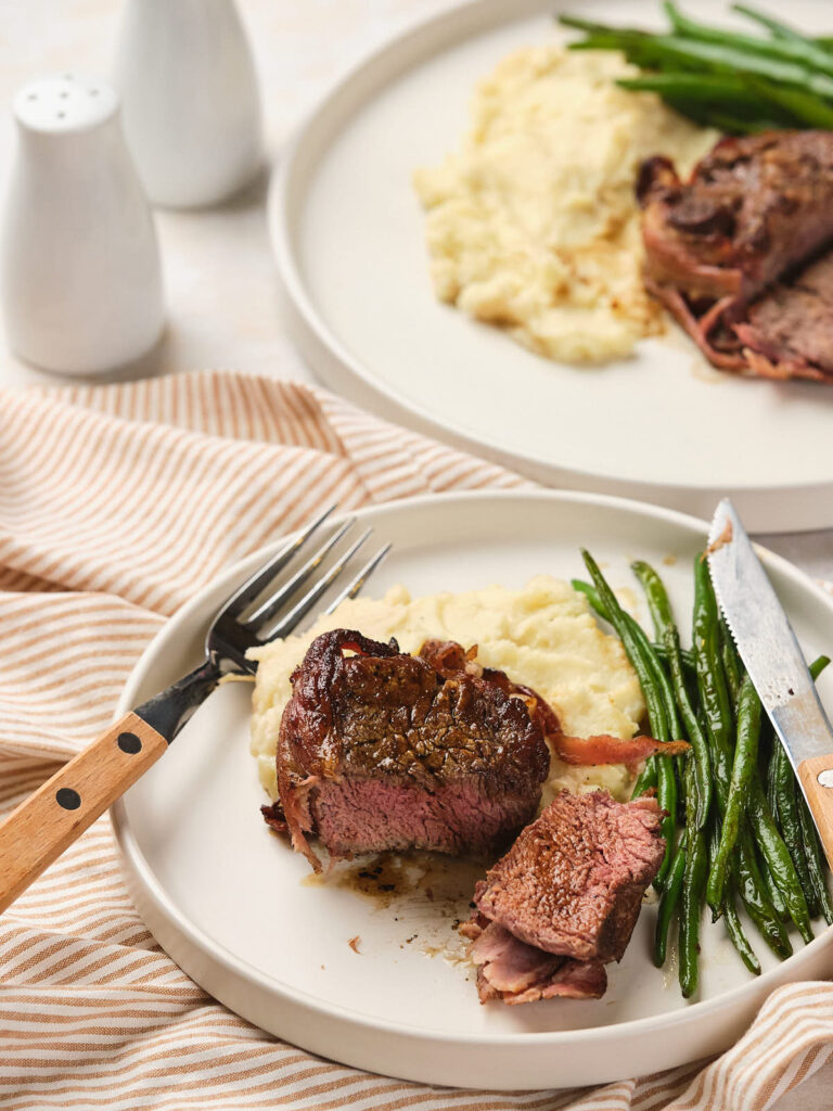 Plate with sliced steak, mashed potatoes, and green beans on a striped cloth.