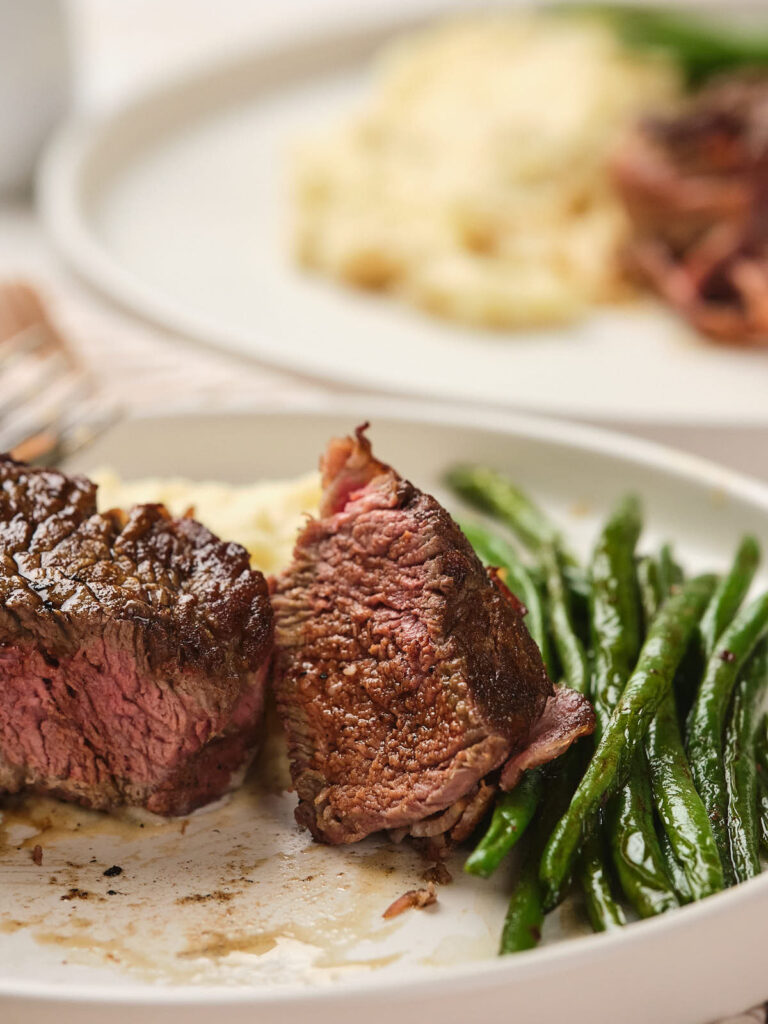 A plate with a sliced piece of cooked steak, green beans, and mashed potatoes. Another similar dish is blurred in the background.