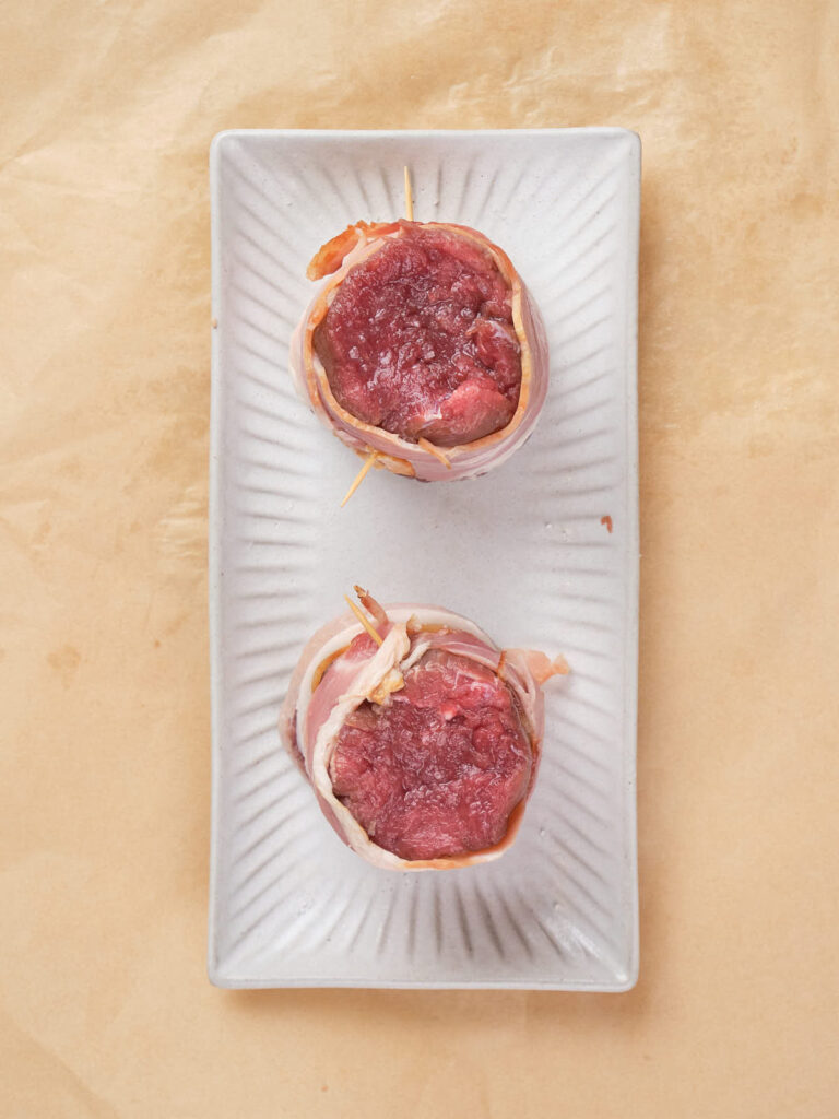 Two raw steaks wrapped in bacon on a rectangular white plate, placed on a beige surface.