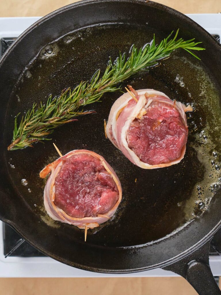 Two bacon-wrapped steak filets and a sprig of rosemary are sizzling in a cast iron skillet.