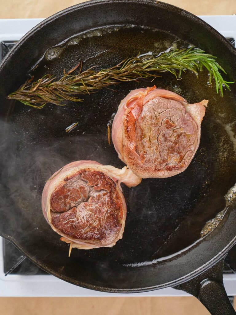 Two bacon-wrapped steaks searing in a black skillet with a sprig of rosemary.