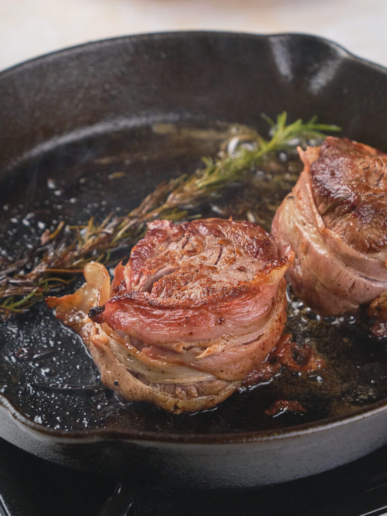 Bacon-wrapped steak cooking in a skillet with a sprig of rosemary.