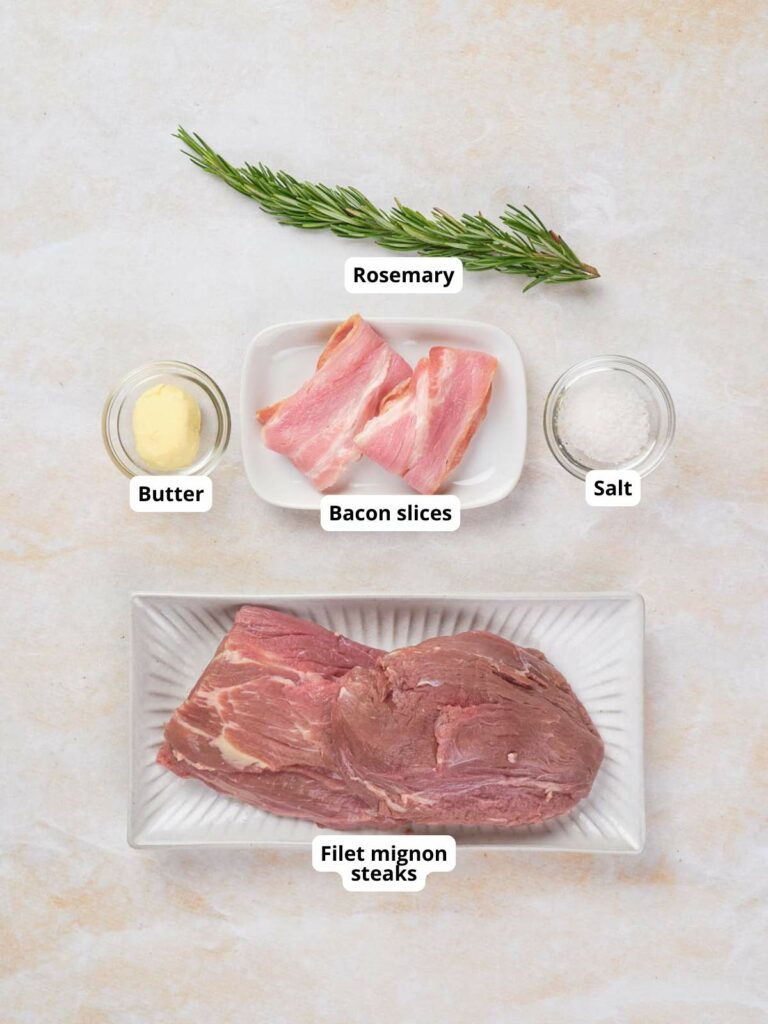 Filet mignon steaks, bacon slices, butter, salt, and rosemary arranged on a light surface.