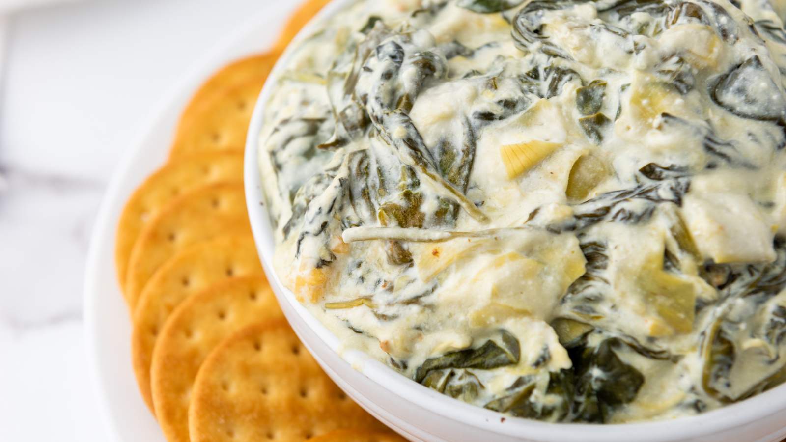 A bowl of creamy spinach artichoke dip with a side of round crackers on a white surface.