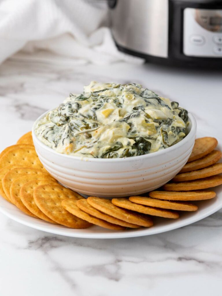 A white bowl of spinach artichoke dip surrounded by round crackers on a plate.