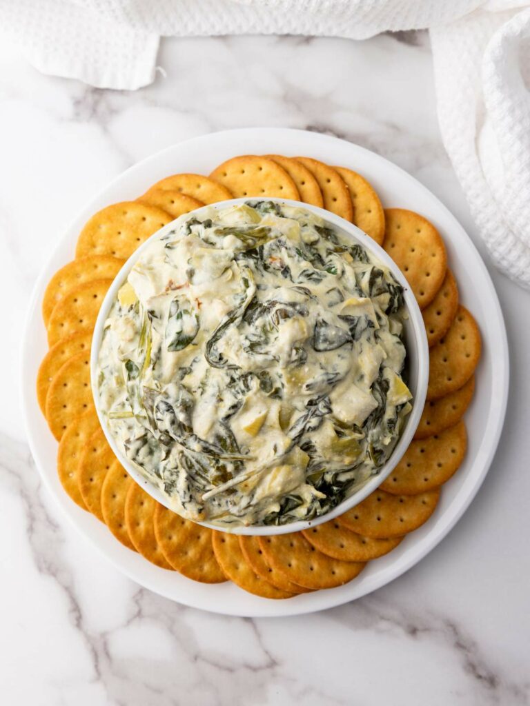 A bowl of creamy spinach artichoke dip is placed on a white plate.