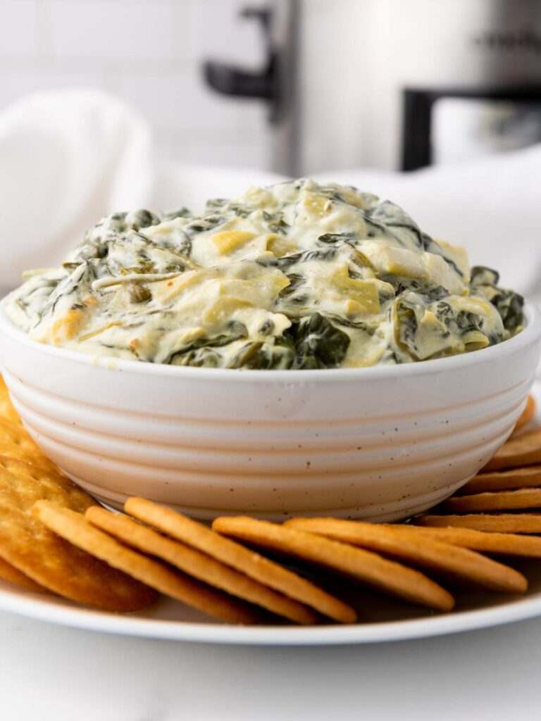 A white bowl filled with creamy spinach artichoke dip, surrounded by a ring of crackers on a plate.