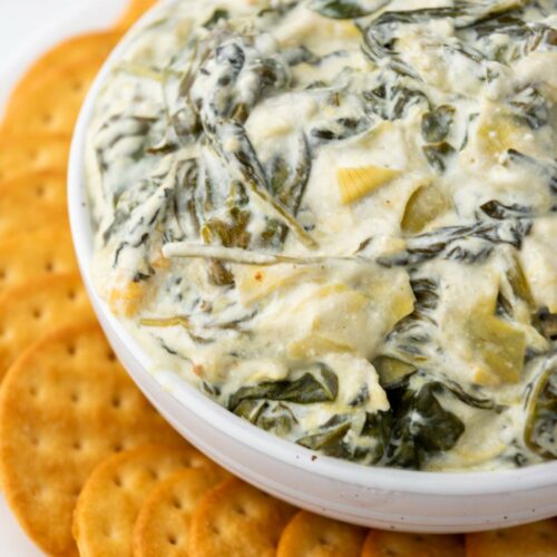 A white bowl filled with creamy spinach and artichoke dip, surrounded by a ring of round crackers on a white plate.