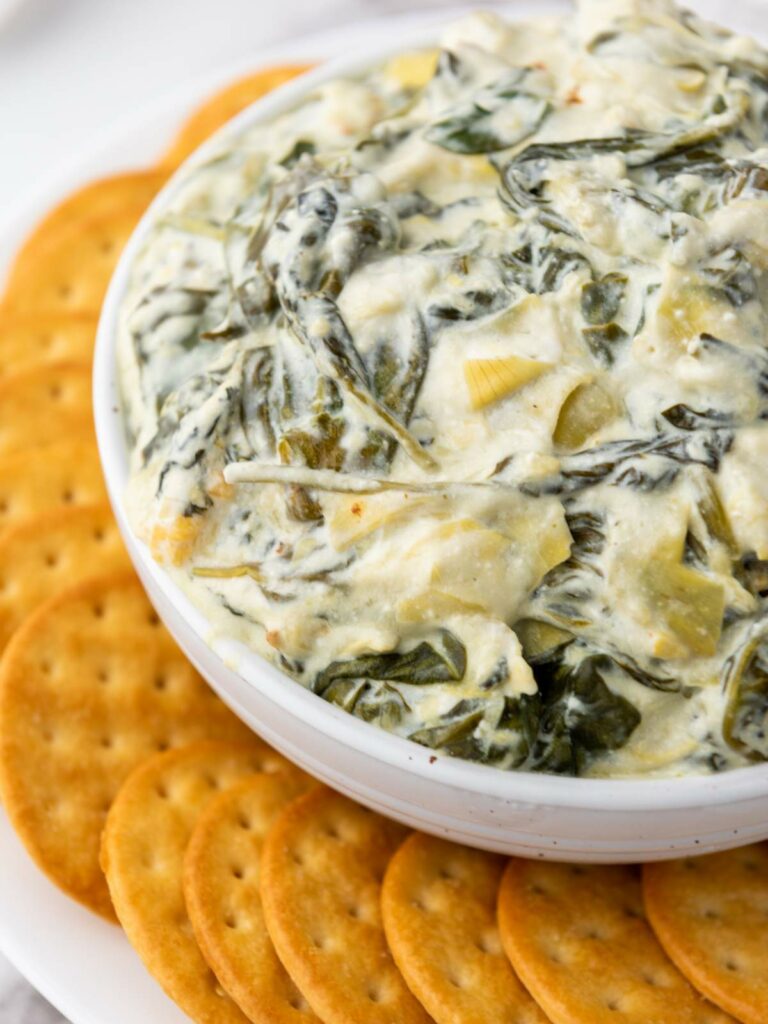 A white bowl filled with creamy spinach and artichoke dip, surrounded by a ring of round crackers on a white plate.