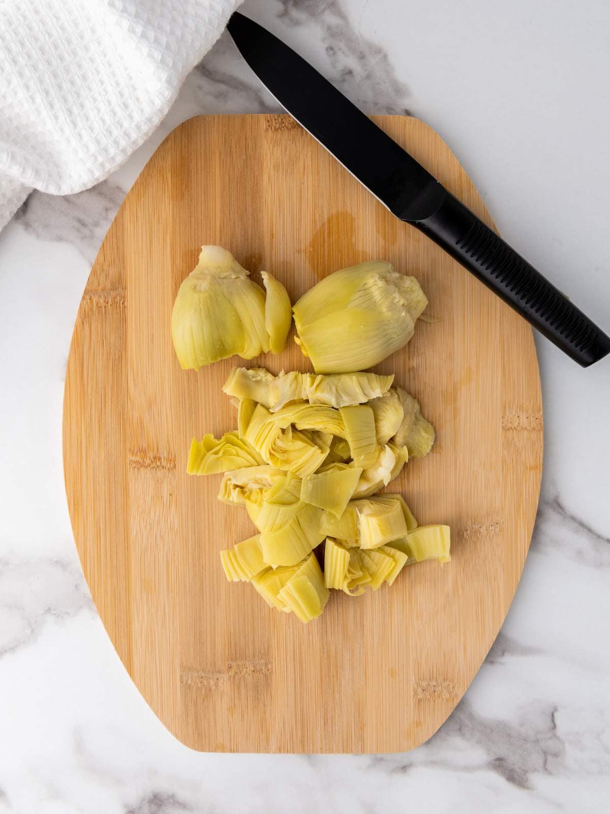 Chopped artichoke hearts on a wooden cutting board with a black knife and white cloth nearby.