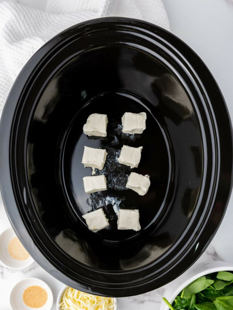 Black slow cooker with eight cubes of cream cheese inside.