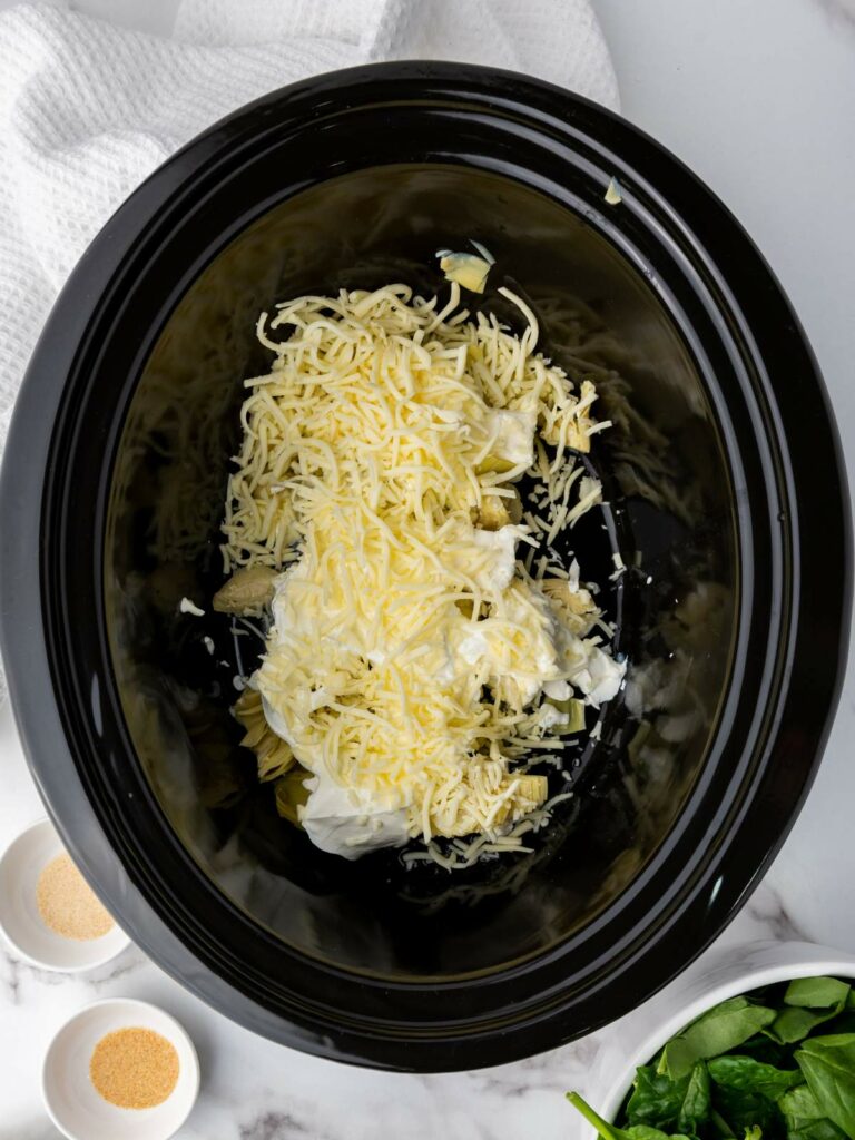 Ingredients in a black slow cooker include shredded cheese, cream, and artichoke hearts.