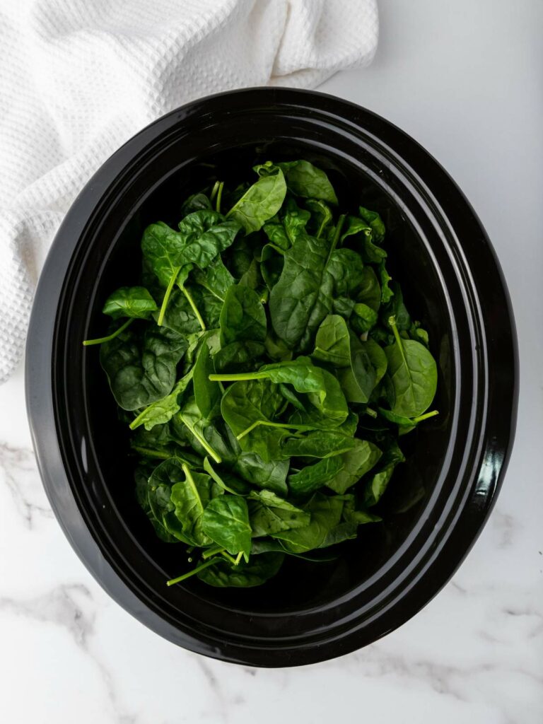 Fresh spinach leaves inside a black slow cooker on a marble surface with a white cloth beside it.