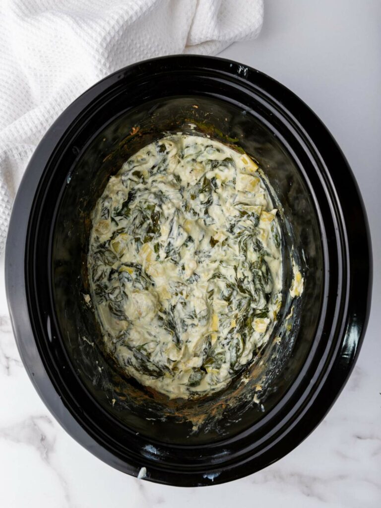 A slow cooker filled with creamy spinach artichoke dip on a marble countertop, partially covered by a white cloth.