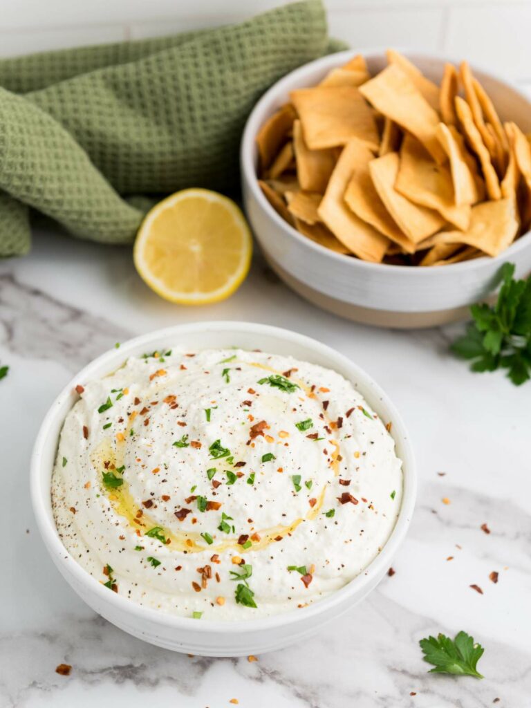 A bowl of creamy feta dip garnished with herbs and spices sits on a marble surface.