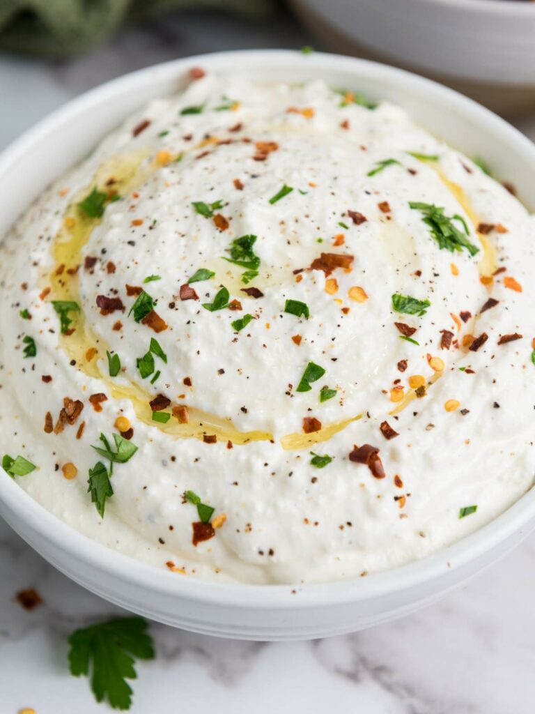 A bowl of feta dip garnished with olive oil, red pepper flakes, and chopped parsley.