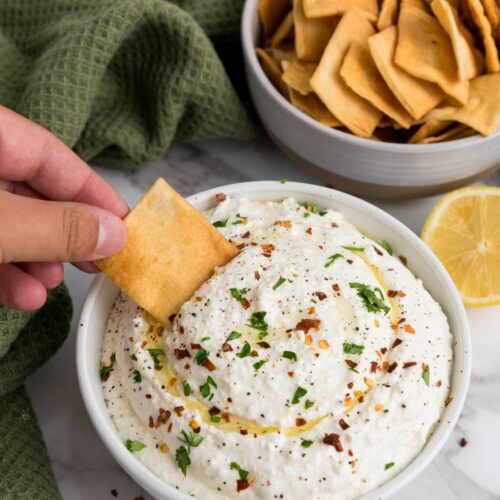 Hand dipping a cracker into a bowl of feta dip topped with herbs and spices.