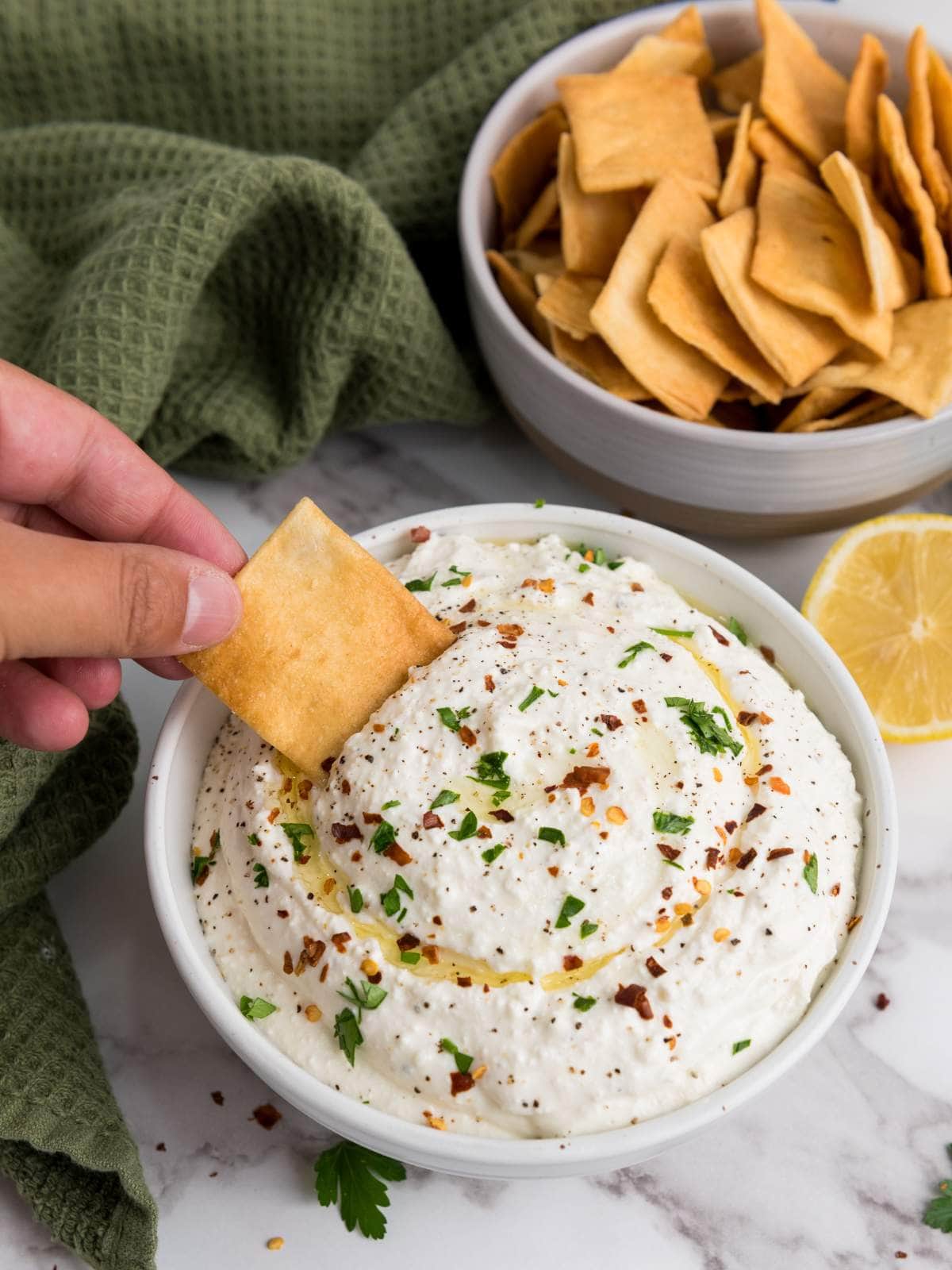 Hand dipping a cracker into a bowl of feta dip topped with herbs and spices.