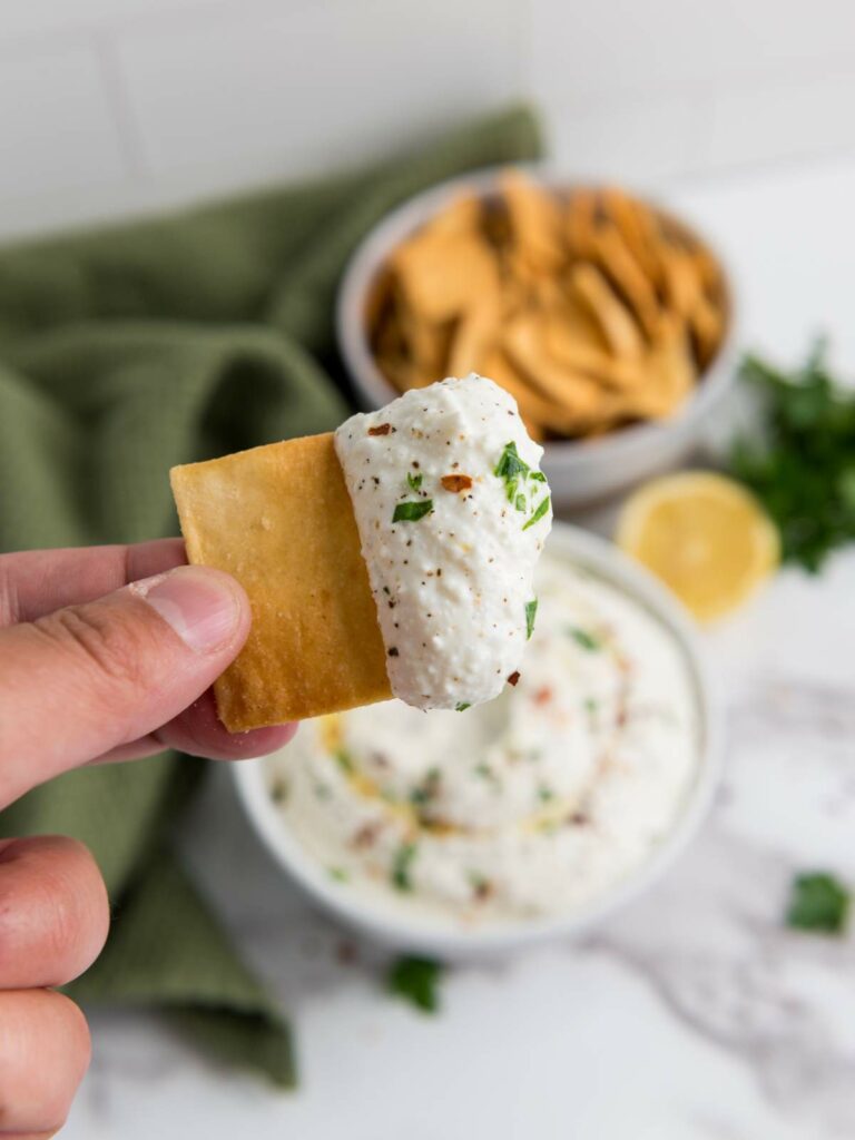 A hand holds a cracker dipped in feta dip with herbs.