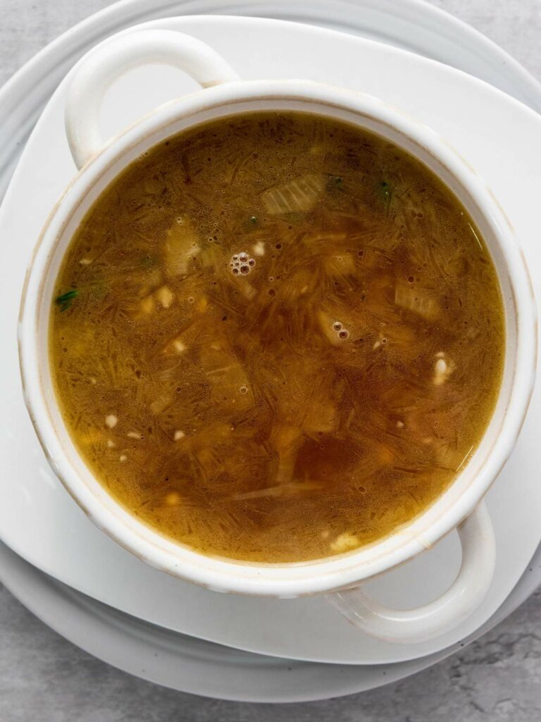 A bowl of clear soup with herbs and small food pieces, served in a white bowl on a matching plate.