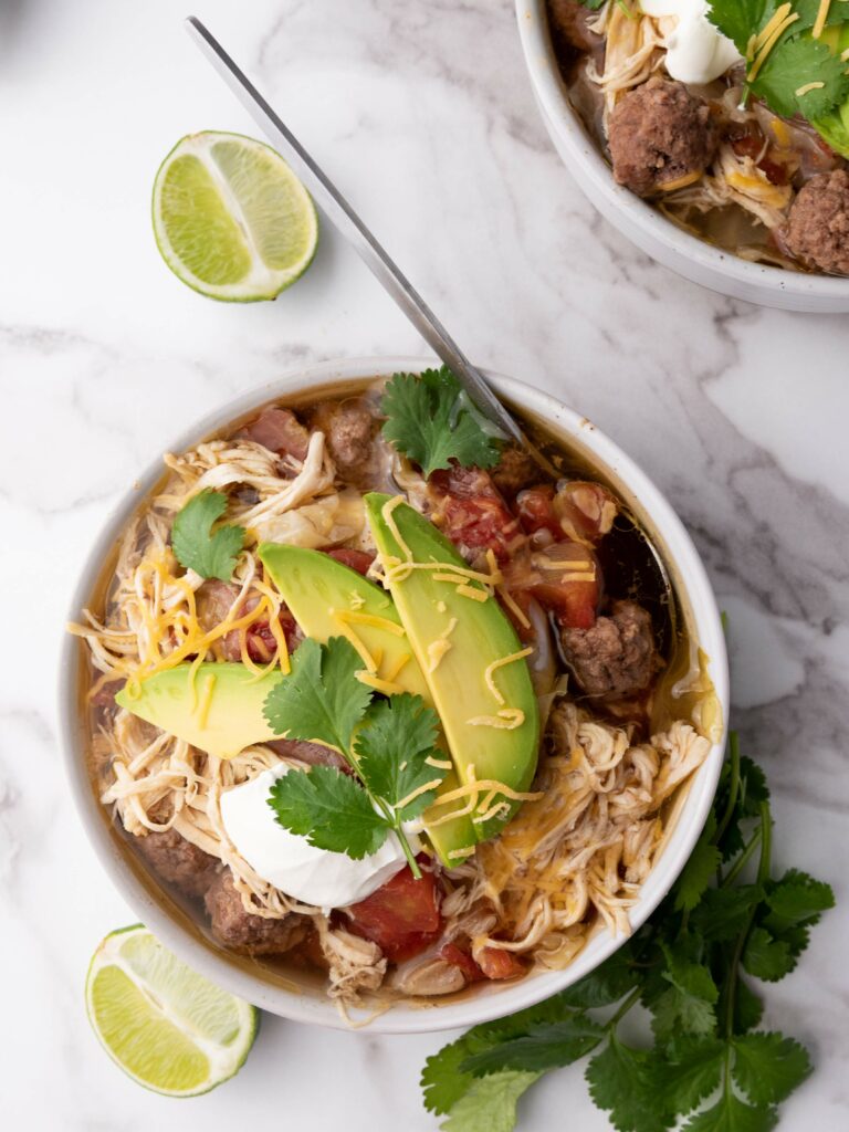 A bowl of taco soup with shredded chicken, avocado slices, cheese, sour cream, and cilantro.