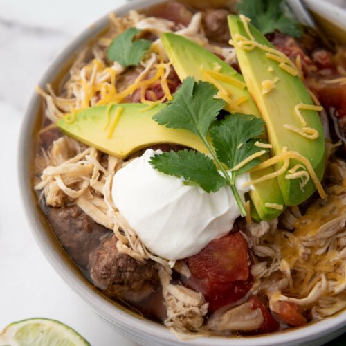 Bowl of taco soup topped with sliced avocado, shredded cheese, sour cream, and cilantro.