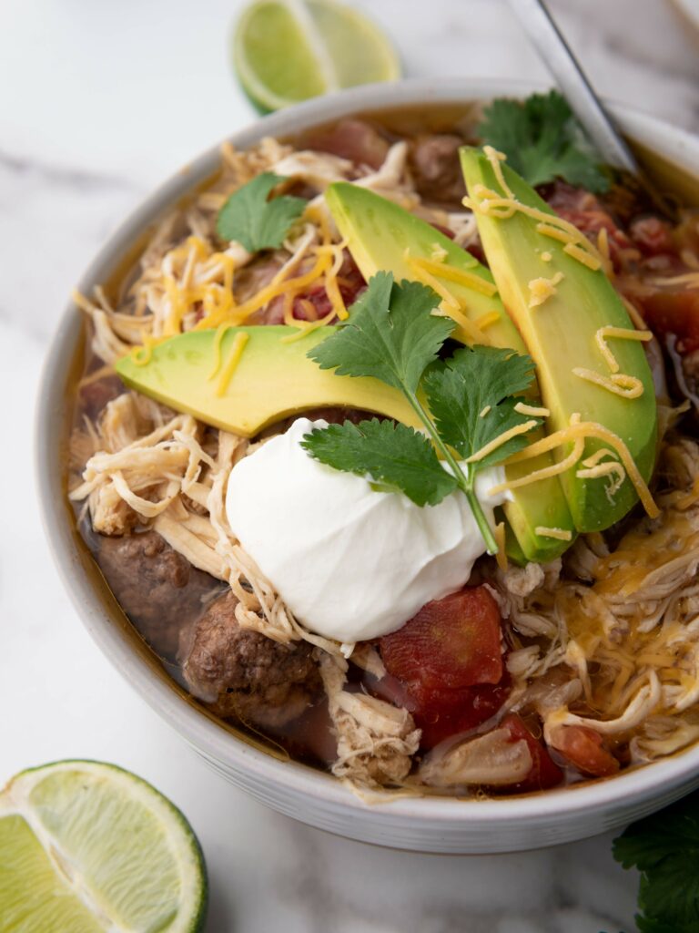 Bowl of taco soup topped with sliced avocado, shredded cheese, sour cream, and cilantro.