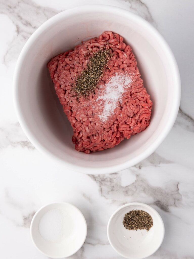 Ground beef in a white bowl with salt and pepper on top.