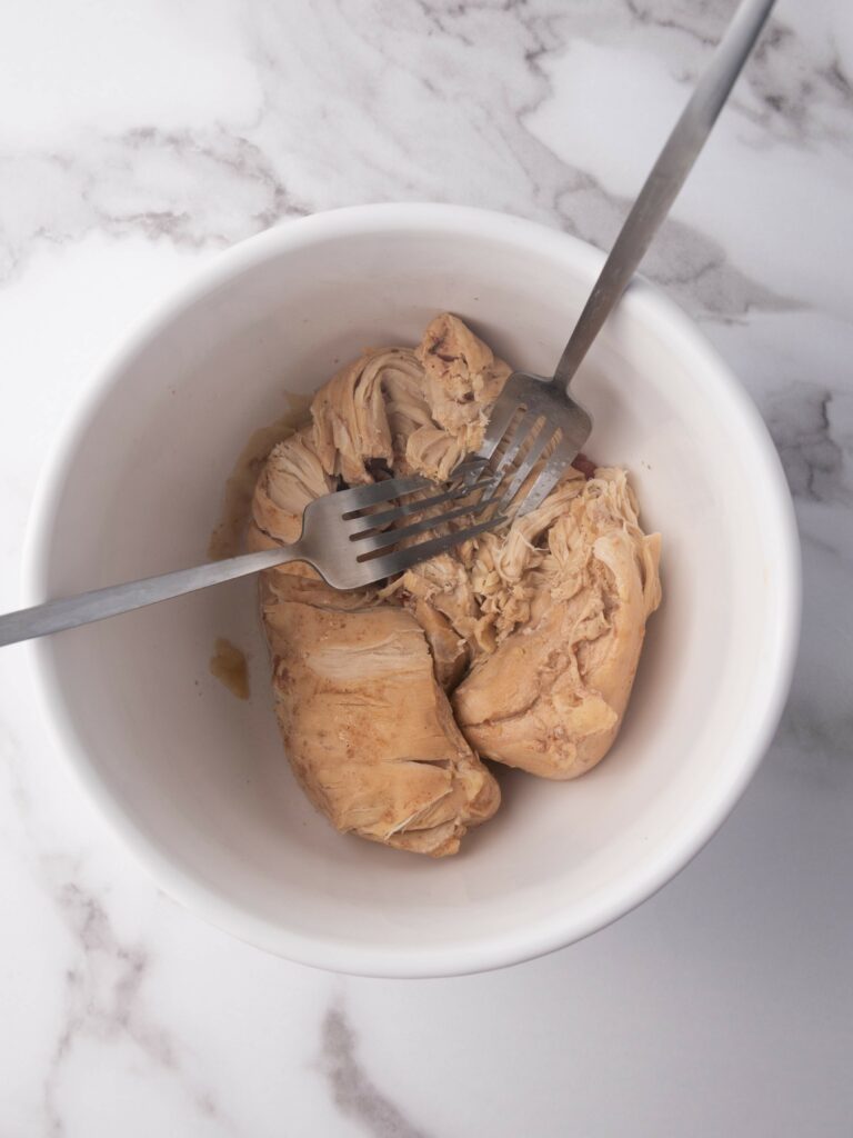 Two forks shredding cooked chicken breasts in a white bowl on a marble countertop.