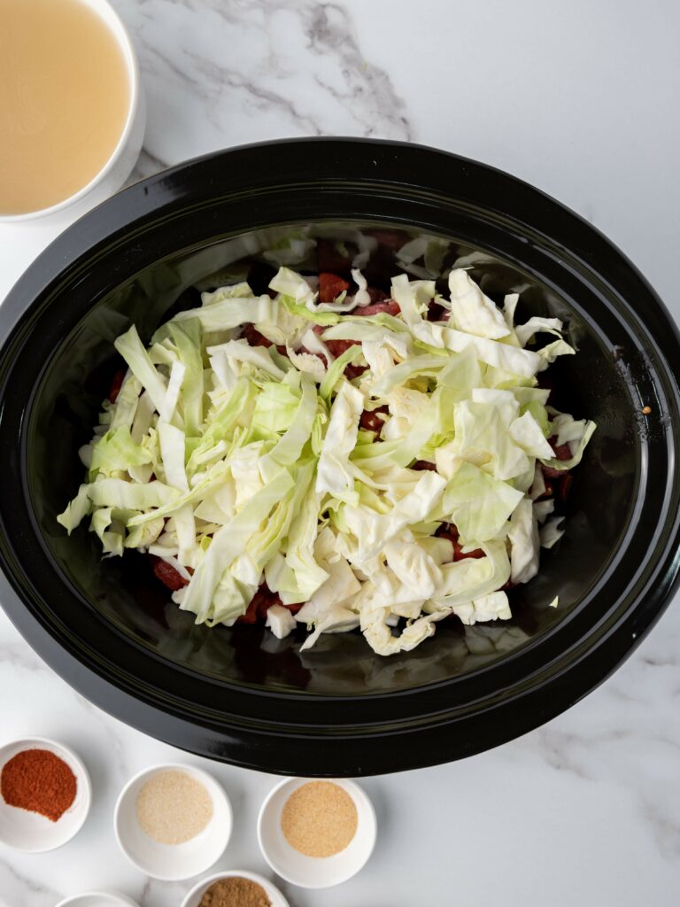 Cabbage in a slow cooker surrounded by bowls containing spices and a cup of broth.