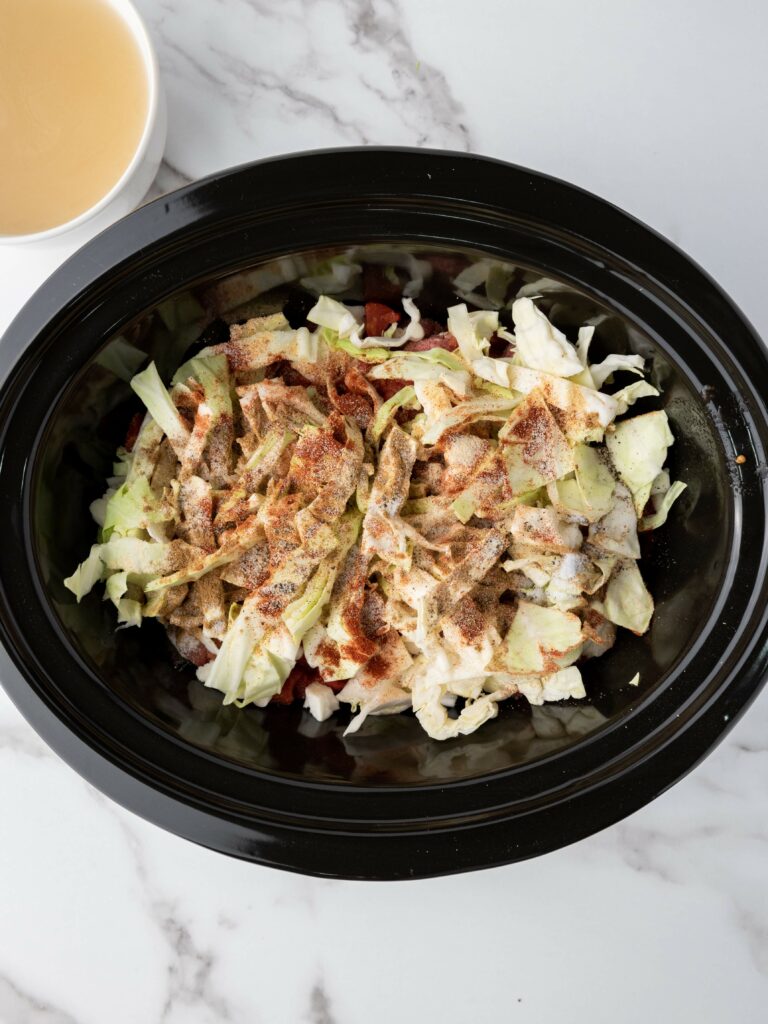 Chopped cabbage and spices in a slow cooker, with a cup of light-colored broth nearby on a marble surface.