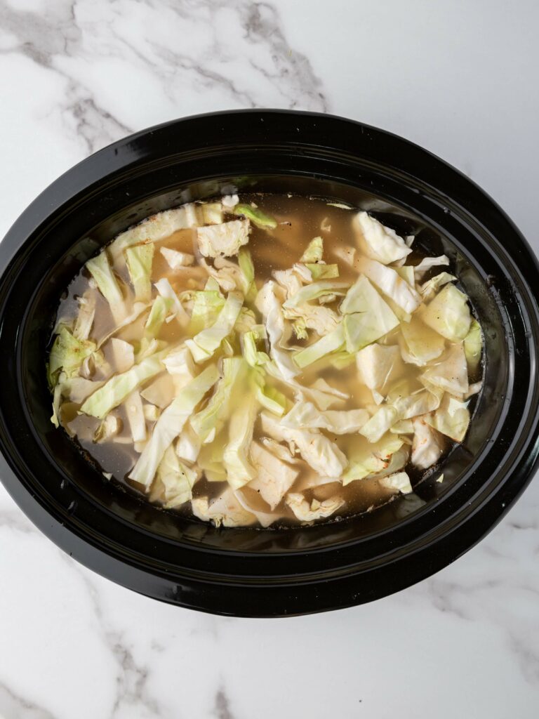 Overhead view of a slow cooker with soup containing cabbage, chicken pieces, and broth on a marble countertop.