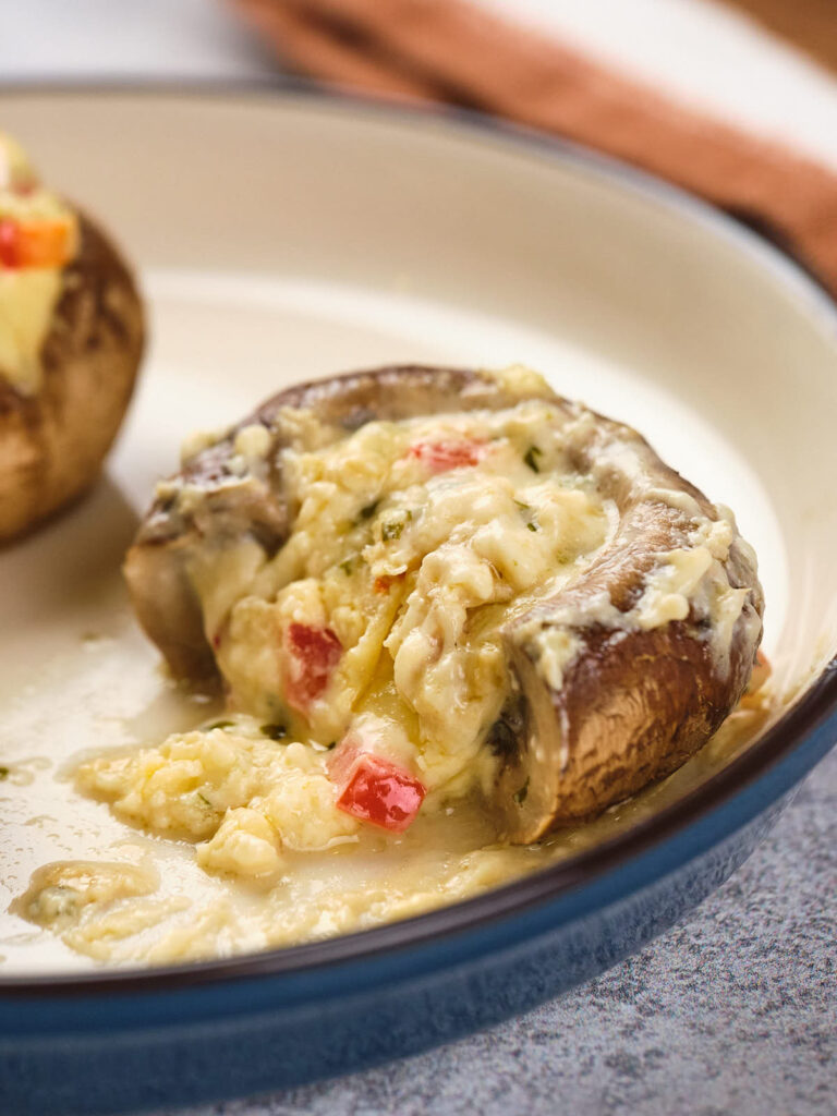 Stuffed mushroom cap filled with melted cheese and small red pepper pieces on a white plate.