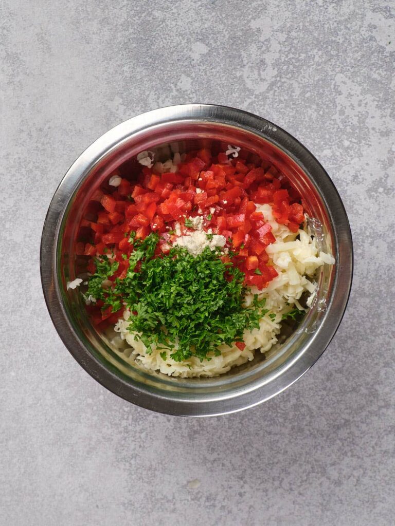 Metal bowl with chopped red peppers, shredded cheese, minced garlic, and parsley.