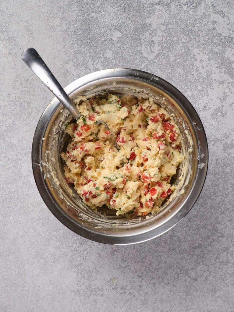 Metal mixing bowl containing dough with red and green bits, a spoon inside.