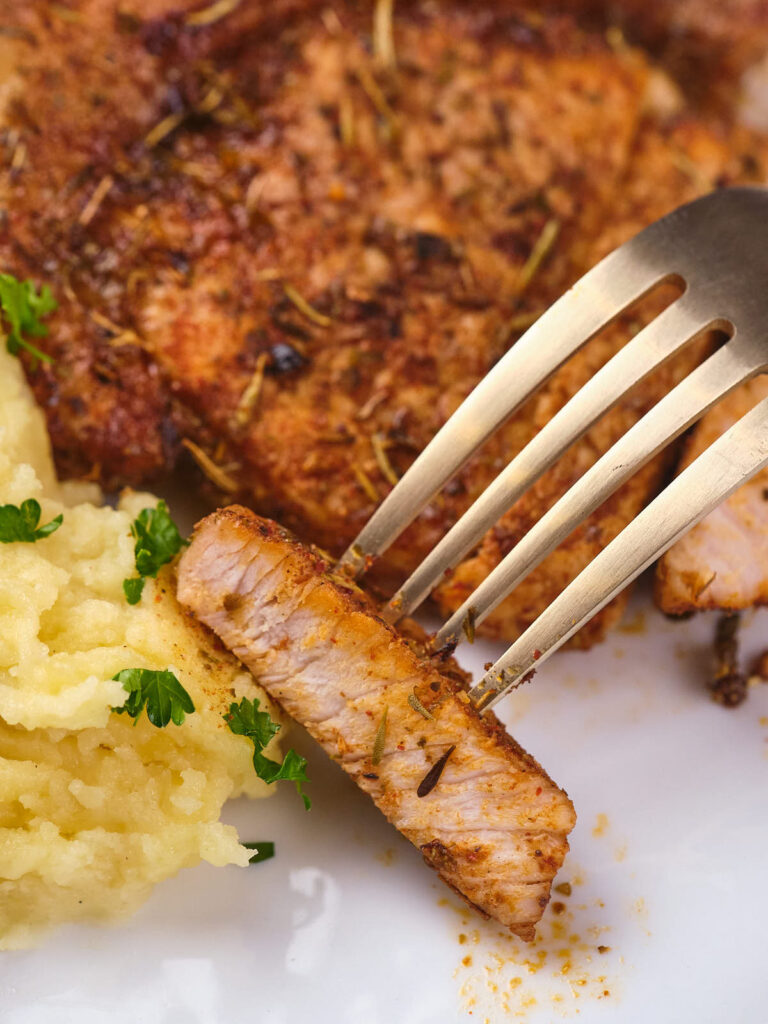 A fork holding a slice of seasoned, cooked boneless pork chops next to mashed potatoes.