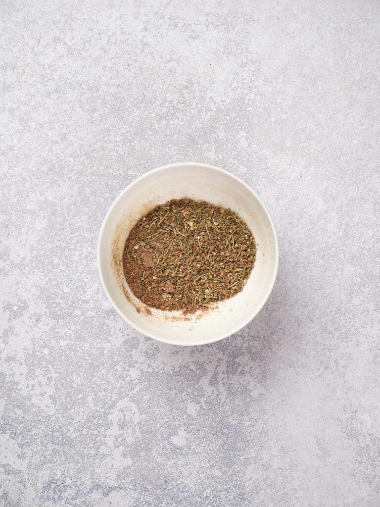 A small bowl containing a mixture of dried herbs and spices on a textured gray surface.