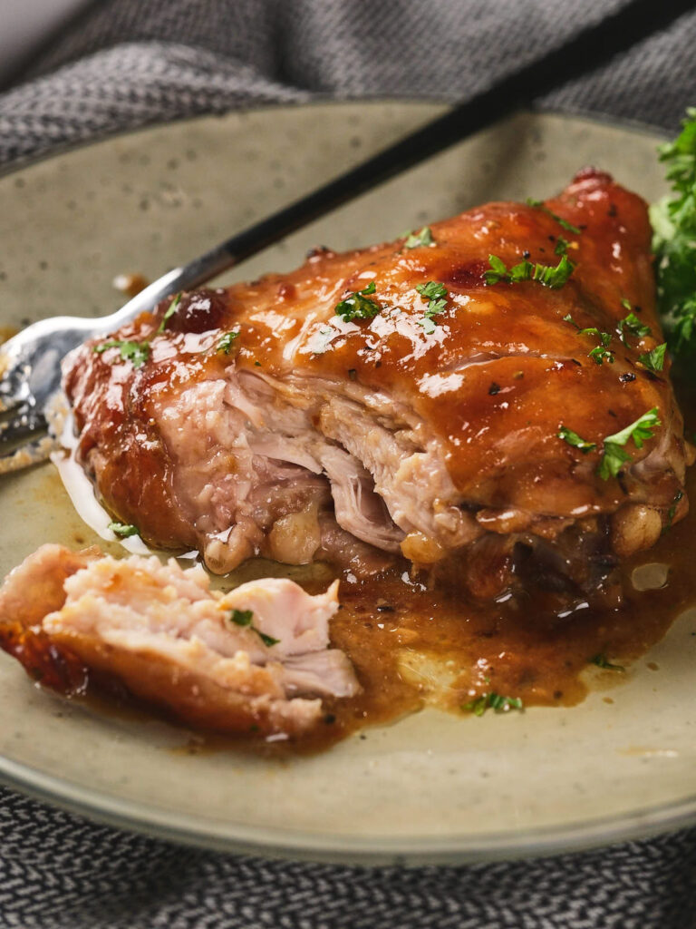 A piece of aptricot glazed cooked chicken partially sliced on a plate, garnished with herbs, with a fork beside it.