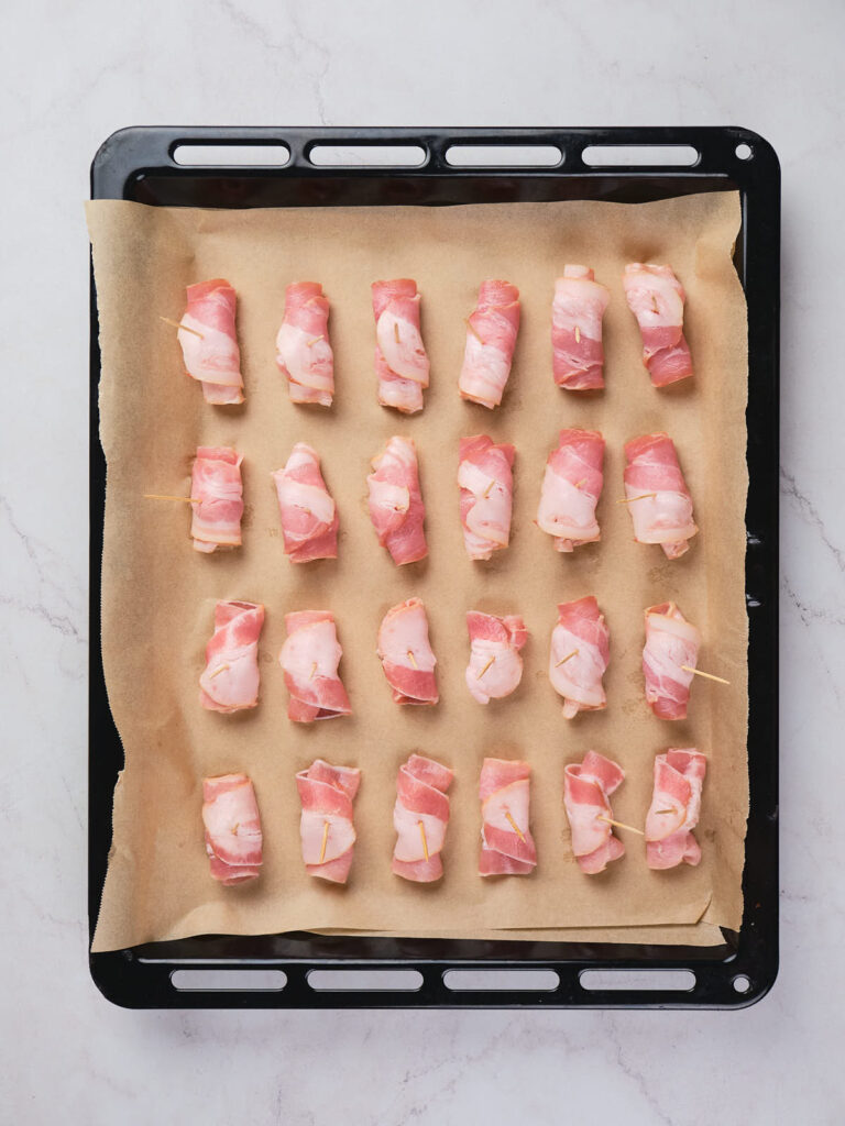 Bacon-wrapped items placed in rows on a parchment-lined baking tray, ready for cooking.