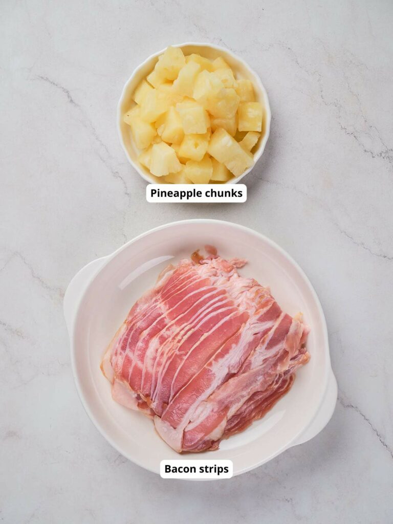 A bowl of pineapple chunks and a dish of uncooked bacon strips on a white marble surface.