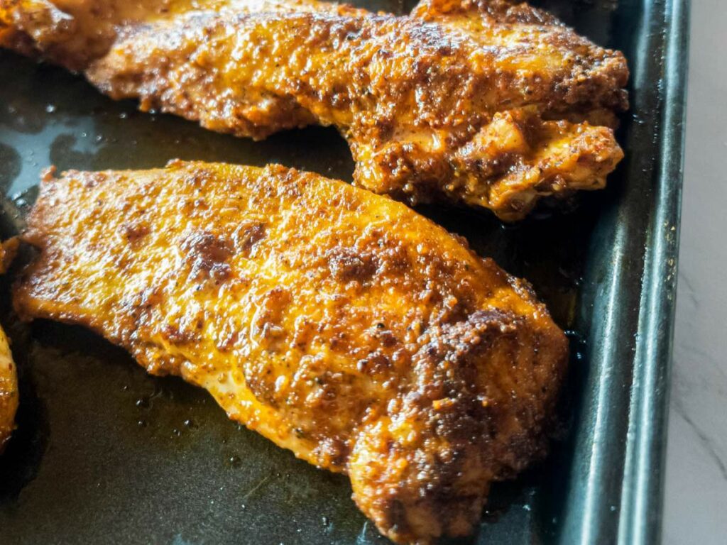 Two seasoned and cooked blackened chicken tenders on a dark tray.