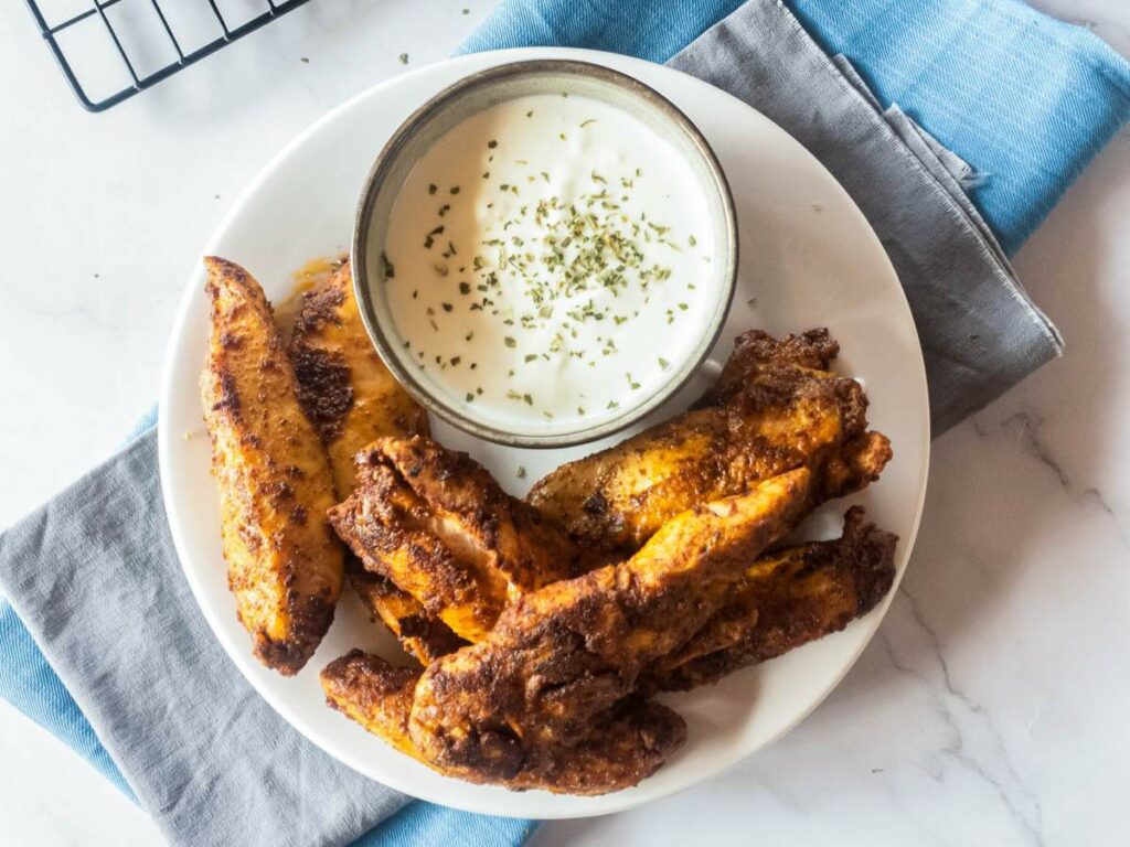 A plate of blackened chicken tenders with a bowl of white dipping sauce topped with herbs.