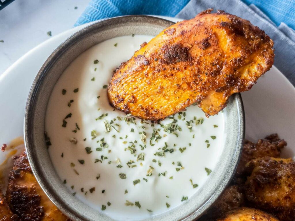 A piece of blackened chicken tender is dipped into a creamy white sauce, garnished with herbs.