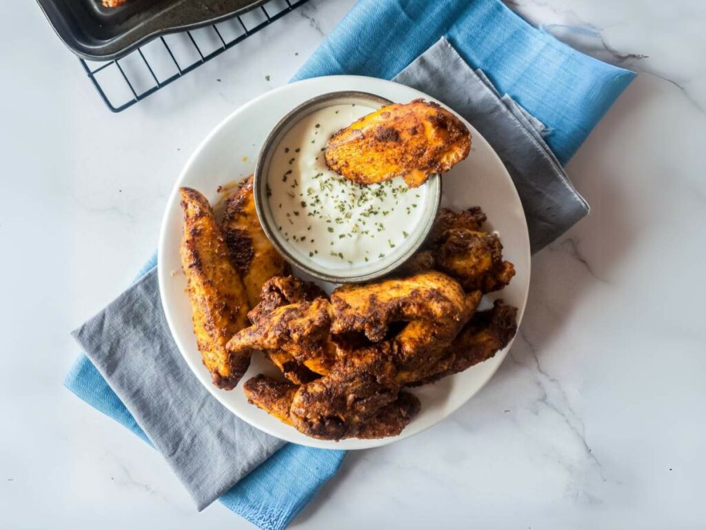 Plate of crispy blackened chicken tenders with a side of creamy dipping sauce, garnished with herbs.