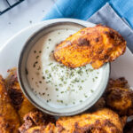 Blackened chicken tenders with a crispy coating, one dipped in a bowl of creamy white sauce garnished with herbs.