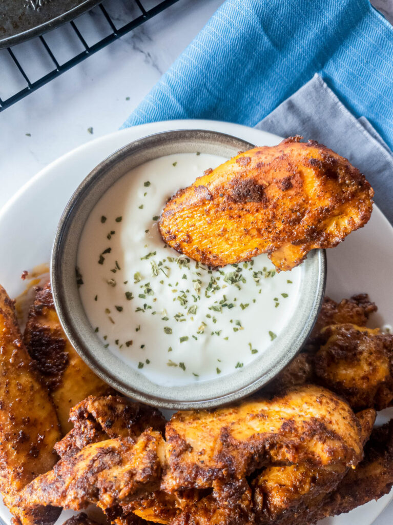 Blackened chicken tenders with a crispy coating, one dipped in a bowl of creamy white sauce garnished with herbs.