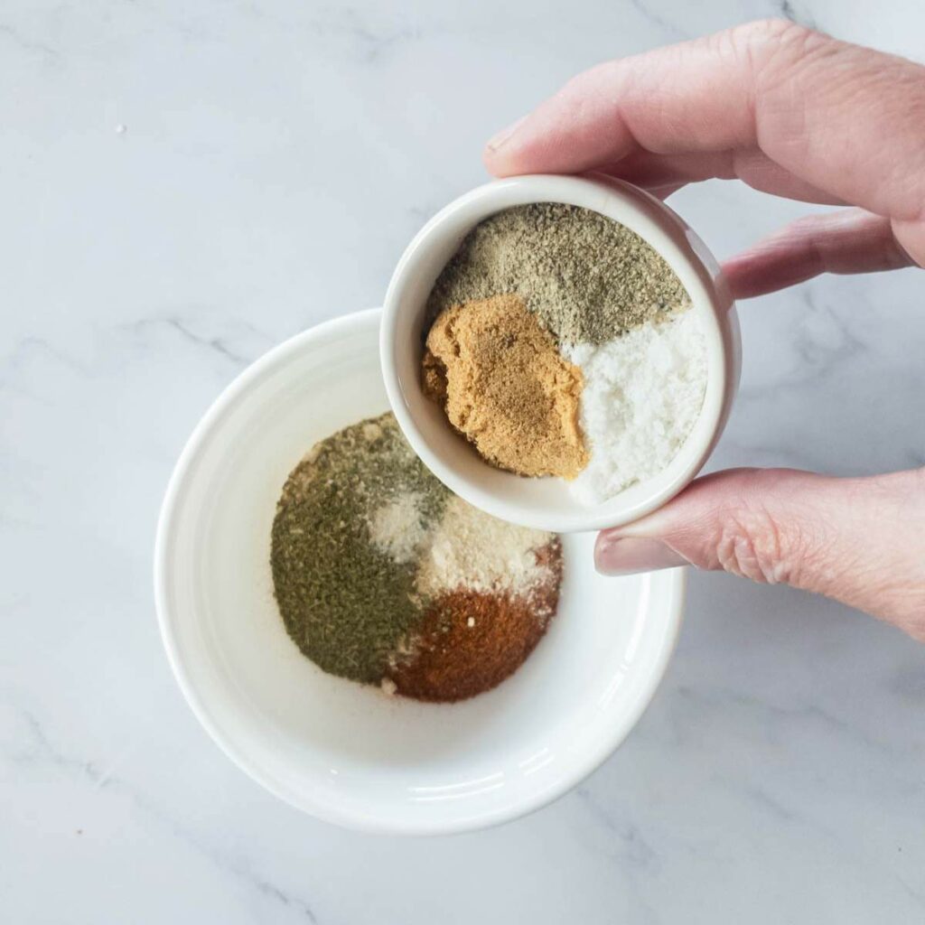 A hand holds a small white bowl containing a mix of spices over a larger bowl with the same spices.